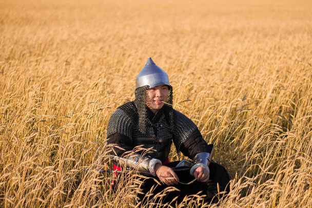 Guerrero asiático con armadura oriental y con un sable en las manos sentado en un campo de trigo
. - Foto, Imagen
