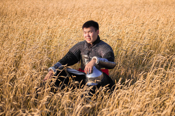 Asiatischer Krieger in orientalischer Rüstung und mit einem Säbel in den Händen, der in einem Weizenfeld sitzt. - Foto, Bild