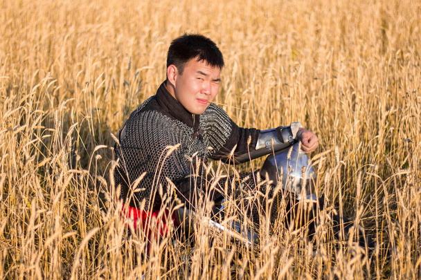 Guerreiro asiático em armadura oriental e com um sabre em suas mãos sentado em um campo de trigo
. - Foto, Imagem