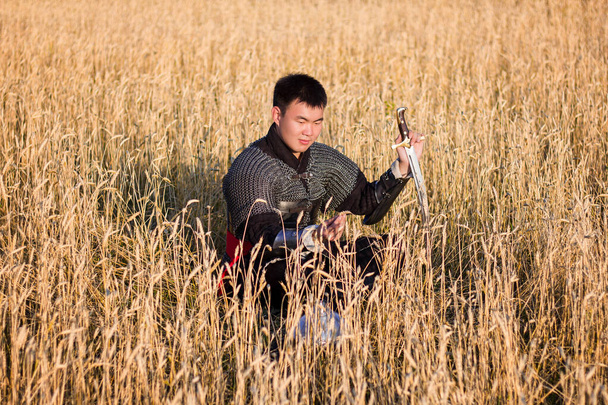 Aziatische krijger in oosterse harnas en met een sabel in zijn handen zittend in een tarweveld. - Foto, afbeelding