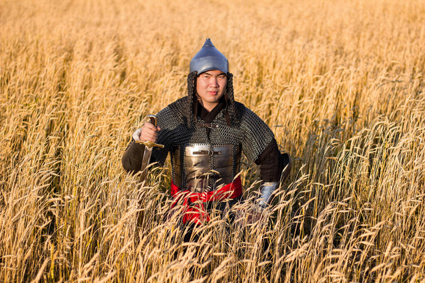 Guerrier asiatique en armure orientale et avec un sabre dans les mains assis dans un champ de blé
. - Photo, image