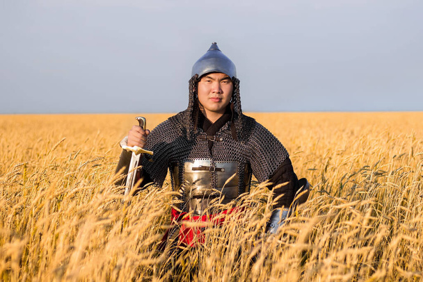 Asian warrior in oriental armor and with a saber in his hands sitting in a wheat field. - Фото, изображение