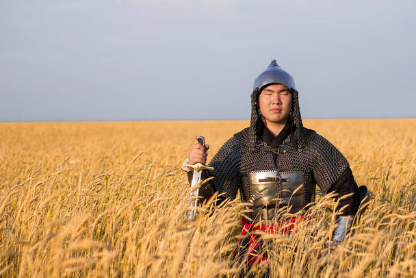 Asian warrior in oriental armor and with a saber in his hands sitting in a wheat field. - Φωτογραφία, εικόνα