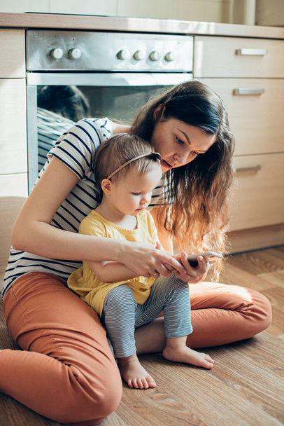 Calm lady sitting on the floor and holding a smartphone while her toddler daughter looking at it - Фото, изображение