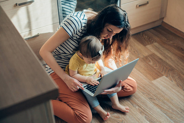 Young woman sitting on the floor and trying to work on the laptop while her child interrupting her stock photo - Φωτογραφία, εικόνα