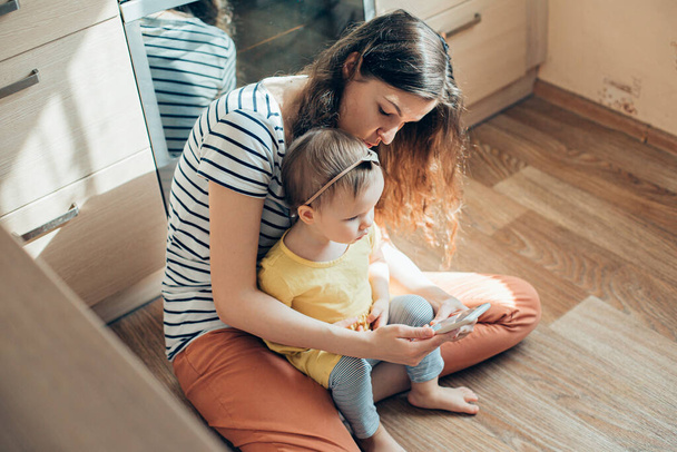 Young lady in casual clothes sitting on the floor with her serious toddler child and holding a smartphone - Fotó, kép
