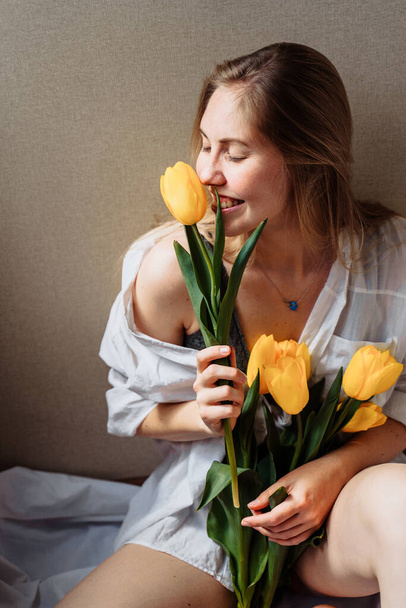 Vertical photo. Attractive blonde girl in lingerie and white shirt sitting in sunshine. Holding yellow tulips, sniffing them, hiding behind flowers, enjoying the sun and life, smiling, laughing - Photo, image