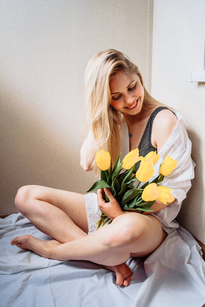 Verticale foto. Aantrekkelijk blond meisje in lingerie en wit shirt zittend in de zon. Gele tulpen vasthouden, ruiken, zich achter bloemen verschuilen, genieten van de zon en het leven, glimlachen, lachen - Foto, afbeelding