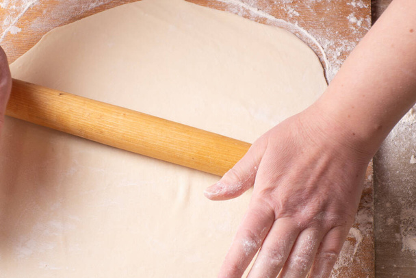 Kesme tahtasının üzerinde merdaneyle hamuru yuvarlayan kadın elleri. Evde yemek pişirmek. - Fotoğraf, Görsel