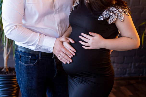 A young couple expecting a baby.A pregnant girl in a black dress with silver wings and her husband in white shirt hug and hold,touche the abdomen.Maternity,preparation,expectation concept.Close up - Valokuva, kuva