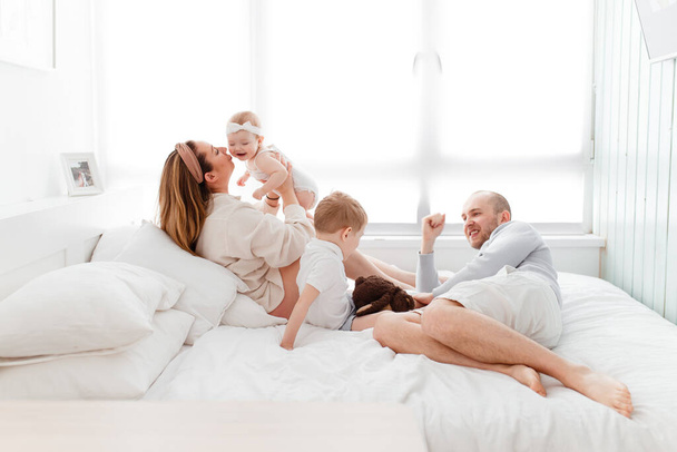 A young happy family with young children lying around, hugging, laughing on the bed in home clothes in a bright cozy house, mom, dad, son and daughter baby - Photo, Image