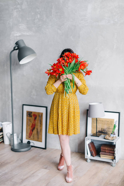 Young woman in a yellow dress hides her face behind flowers. Girl with big bouquet of red tulip flowers staying home. Fashion portrait in the stylish interior - Zdjęcie, obraz