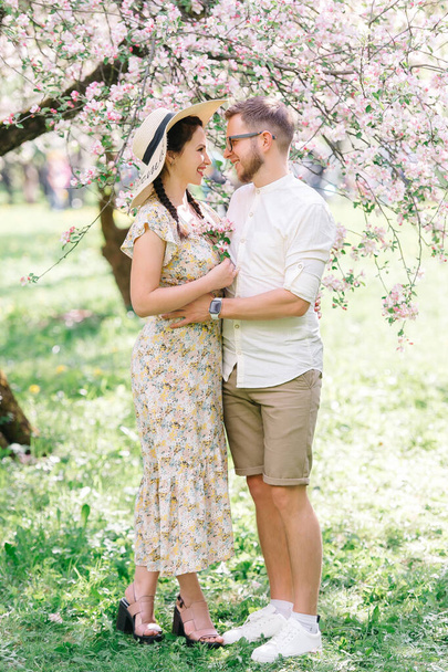 A young couple on a date in a flowering garden in springtime. Love story under the sakura blossom tree. Summer fashion concept: floral dress and straw hat - Foto, immagini
