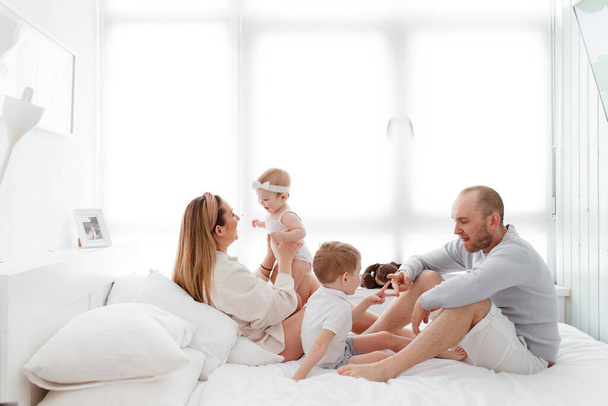A young happy family with young children lying around, hugging, laughing on the bed in home clothes in a bright cozy house, mom, dad, son and daughter baby - Photo, Image