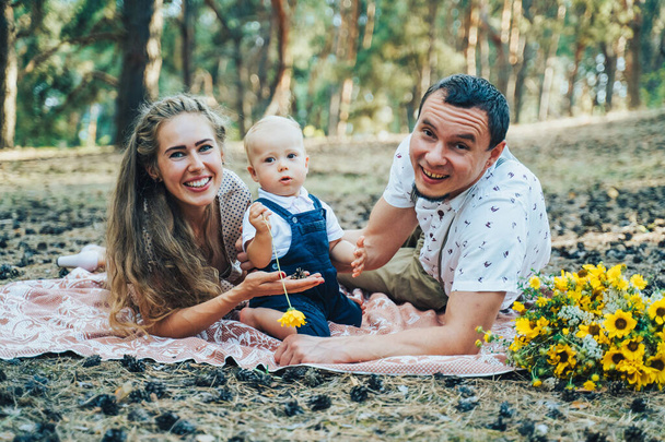 Young attractive parents on vacation in a rural forest. They like trips out of town very much. Sitting and lying with their little child boy on the bedding cover, looking at the camera and smiling - Foto, Imagem