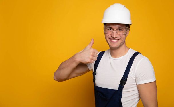 Confident professional. Close-up portrait of attractive man in a working suit, helmet and transparent glasses, who is looking in the camera and showing thumbs up. - Fotó, kép