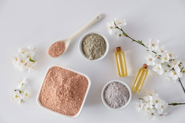 Ceramic bowl with red clay powder, ingredients for homemade facial and body mask or scrub and fresh sprig of flowering cherry on white background. Spa and bodycare concept. - Photo, Image