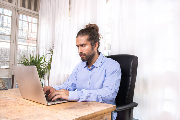άντρας με μακριά μαλλιά που δουλεύει με το λάπτοπ του σε ένα γραφείο - Φωτογραφία, εικόνα