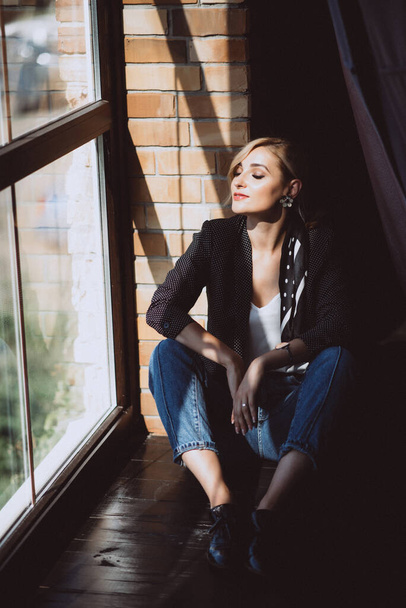Beautiful young woman blonde in jeans with a polka dot jacket and a scarf in her hair in a loft studio near the window with sunbeams. Soft selective focus. - Фото, изображение