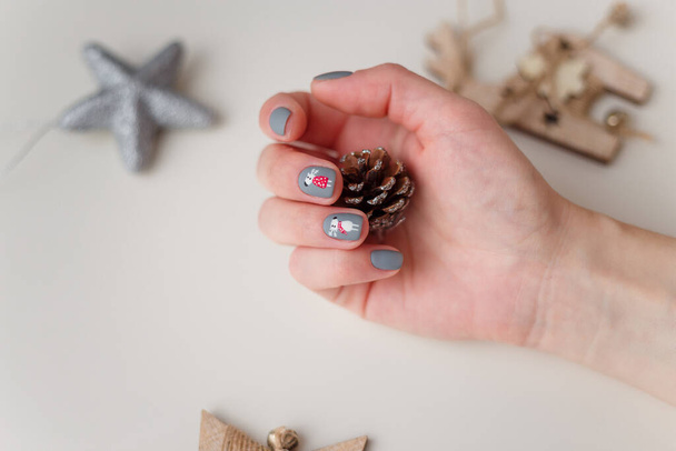Female hands with Merry Christmas reindeer manicure on finger nails holding decorative pine cone. White background with silver and wooden toys around. New Year, nails care, beauty and fashion concept. - Photo, Image