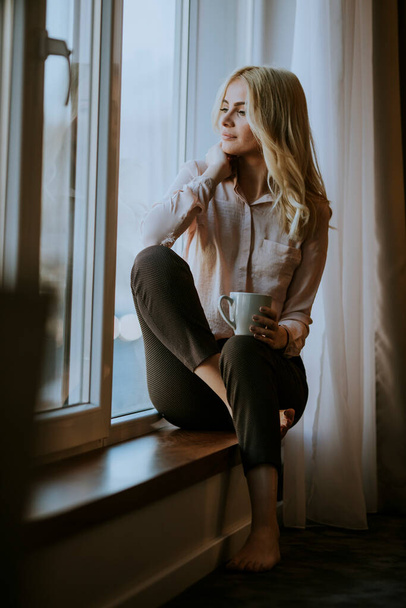 Mooie jonge vrouw met kopje thee of koffie zittend en drinkend op de vensterbank thuis - Foto, afbeelding