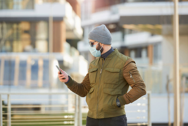 居心地の良い通りでスマートフォンをチェックする拡散コロナウイルスを避けるために、医療用フェイスマスクの男。横になって立っている男は、 COVID 19に対して灰色のキャップ、サングラス、顔マスクを身に着けています. - 写真・画像