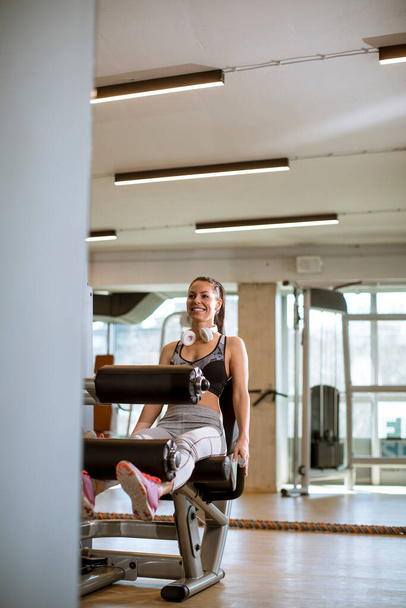 Красивая молодая женщина с упражнениями на растяжение ног и кудрявый автомат в тренажерном зале - Фото, изображение