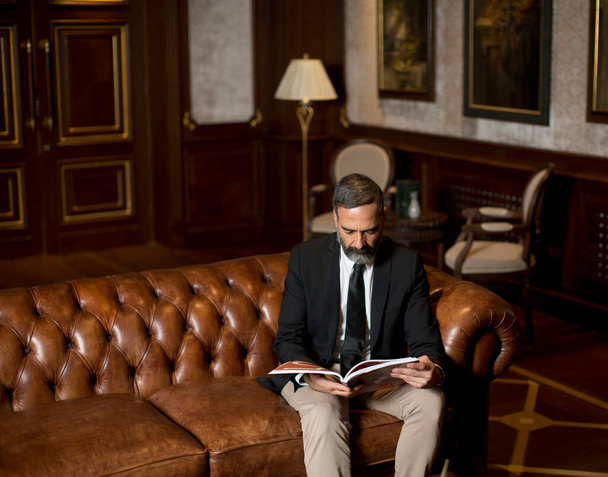 Handsome mature man in elegant suit reading a book in a luxurious apartment - Фото, зображення