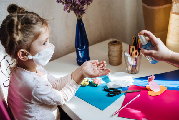 母親は消毒剤の抗菌スプレーを少女の手に吹きかける。Covid-19感染に対する予防措置。自宅で隔離された保護マスクの病気の子供. - 写真・画像
