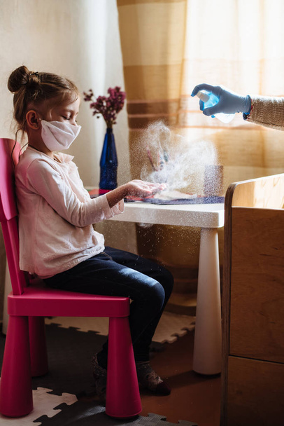 Mom sprays a disinfectant antibacterial spray on the girl's hands. Preventive measures against Covid-19 infection. Sick child in protective mask on isolation at home. - Photo, Image