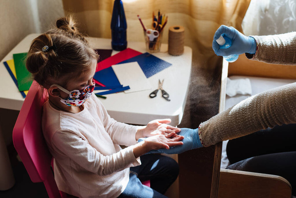 Mom sprays a disinfectant antibacterial spray on the girl's hands. Preventive measures against Covid-19 infection. Sick child in protective mask on isolation at home. - 写真・画像