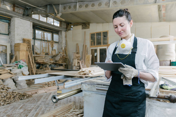 Confident woman working as carpenter in her own woodshop. She using a tablet pc and writes notes while being in her workspace. Small business concept. - Zdjęcie, obraz