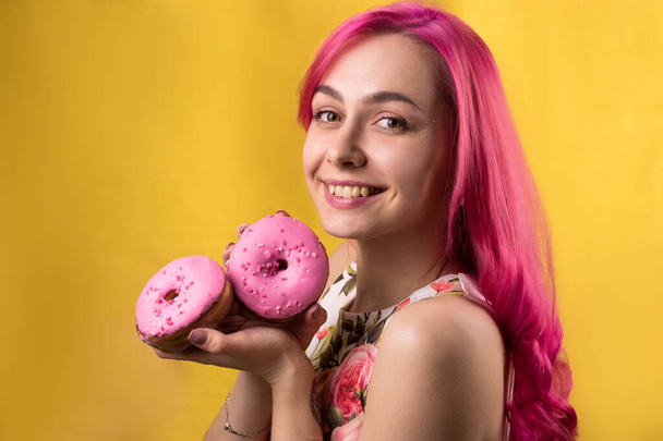 mooi meisje met roze haar houdt een donut. tevreden lachende tiener op een gele achtergrond close-up. - Foto, afbeelding