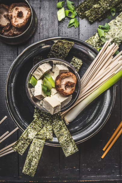 Aziatische soepbereiding in kom met udon noedels, tofu, shiitake champignons, nori en groene ui op donkere rustieke achtergrond, bovenaanzicht - Foto, afbeelding