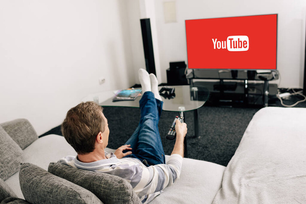 KYIV, UKRAINE - APRIL 14, 2020: man in jeans holding remote controller near tv screen with youtube at home  - Foto, immagini