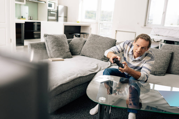 KYIV, UKRAINE - APRIL 14, 2020: selective focus of man playing video game and holding joystick near smartphone with white screen on coffee table  - Foto, Imagen