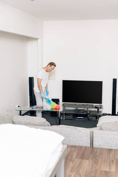 selektiver Fokus eines fröhlichen Mannes mit Staubwedel beim Putzen des Wohnzimmertisches  - Foto, Bild