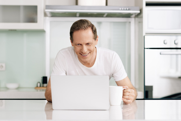 веселый фрилансер смотрит на ноутбук и держит чашку дома
 - Фото, изображение