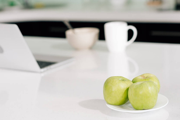 foco seletivo de maçãs verdes perto de laptop, tigela e xícara na mesa
  - Foto, Imagem
