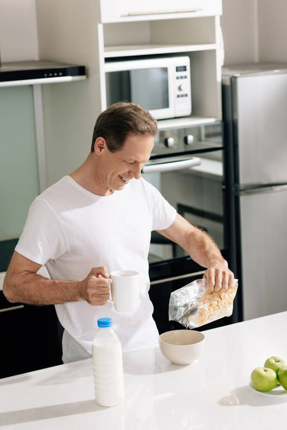 Lächelnder Mann gießt Cornflakes in Schüssel neben Flasche mit Milch und Äpfeln - Foto, Bild
