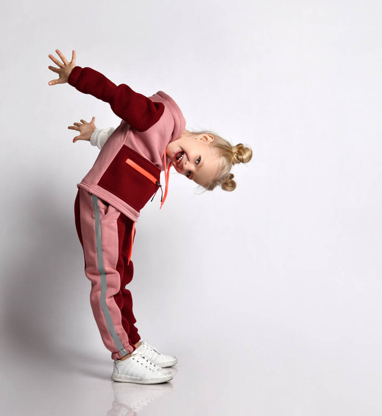 Laughing blonde schoolgirl in pink and red jym suit leaning head forward taking arms back. Full length portrait isolated on light grey - Foto, immagini