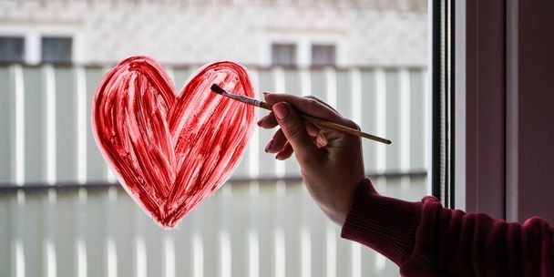 handen van een meisje met een penseel schilderen rood hart op een raam, kopieer ruimte voor tekst, blijf thuis, quarantaine vrije tijd, laten we allemaal goed zijn - Foto, afbeelding