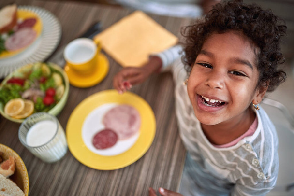top view of a happy smiling face of young african girl at the kitchen table, having breakfast. - Фото, изображение