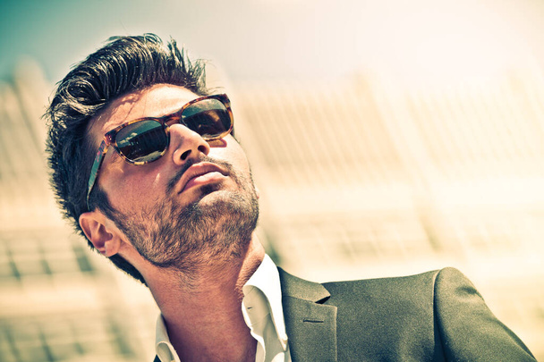 Handsome businessman with sunglasses, outdoor in the city. Charming and modern style, with shirt and suite. Cool hairstyle. Behind him a high-rise office building. - Photo, Image