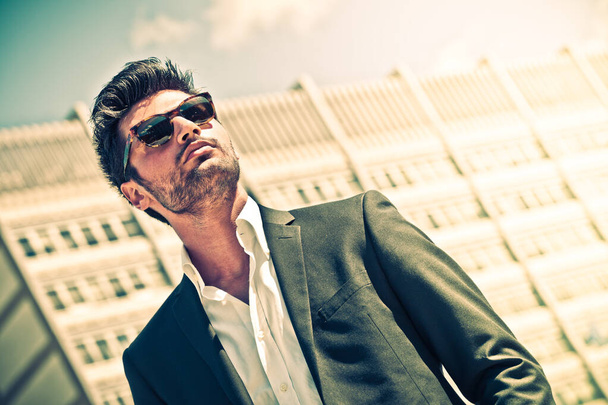 Hombre de negocios guapo con gafas de sol, al aire libre en la ciudad. Encantador y moderno estilo, con camisa y suite. Bonito peinado. Detrás de él un edificio de oficinas. - Foto, imagen