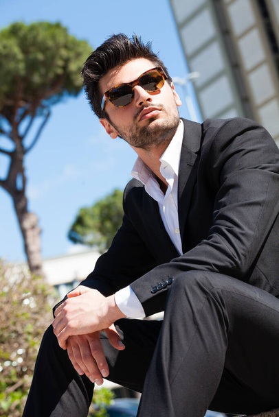Joven y seguro hombre exitoso sentado en la ciudad. Llevando gafas de sol, detrás de él un moderno edificio de oficinas. Pelo moderno, camisa blanca y chaqueta
. - Foto, imagen
