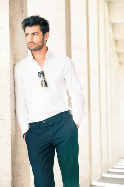 Handsome and stylish young man. Hair and beard fashionable. A beautiful young Italian man is outdoors. He is leaning against a marble wall. He wears a white shirt and dark trousers. Sunglasses. - Fotoğraf, Görsel
