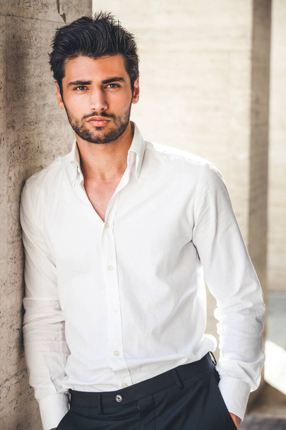 Portrait of young handsome man in white shirt outdoor. Nice appearance with stylish hair and beard. Leaning with a side on a wall - Foto, immagini