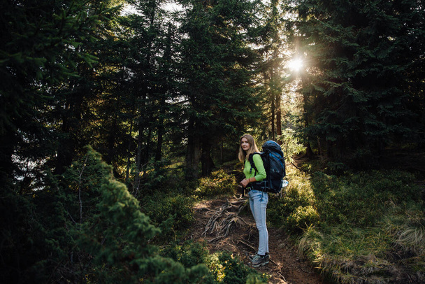 Atrakcyjna turystka z plecakiem patrząca wstecz w kamerę i uśmiechnięta. Słoneczko w lesie. - Zdjęcie, obraz