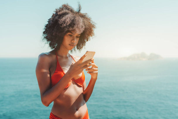 Dazzling young African-American female with curly afro hair standing in a swimsuit on a beach with ocean and a small island in the background and using her smartphone; a copy space place on the right - Foto, Bild